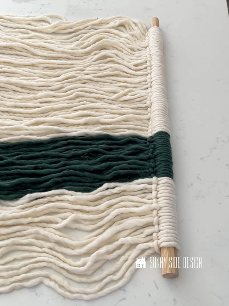 Cream and green yarn are looped onto a dowel with a larks head knot, forming the soon to be fiber wall art.