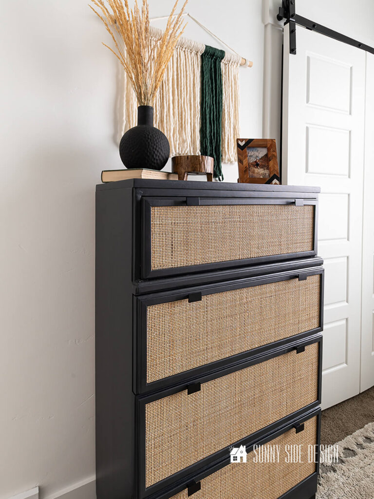 Complete fiber wall art hung on a wall in a bedroom above a black and cane dresser. Dresser decorated with a black vase and grass, footed wooden bowl, and picture frame.