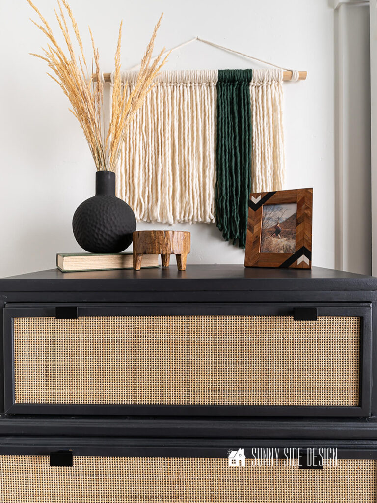 Complete fiber wall art hung on a wall in a bedroom above a black and cane dresser. Dresser decorated with a black vase and grass, footed wooden bowl, and picture frame.