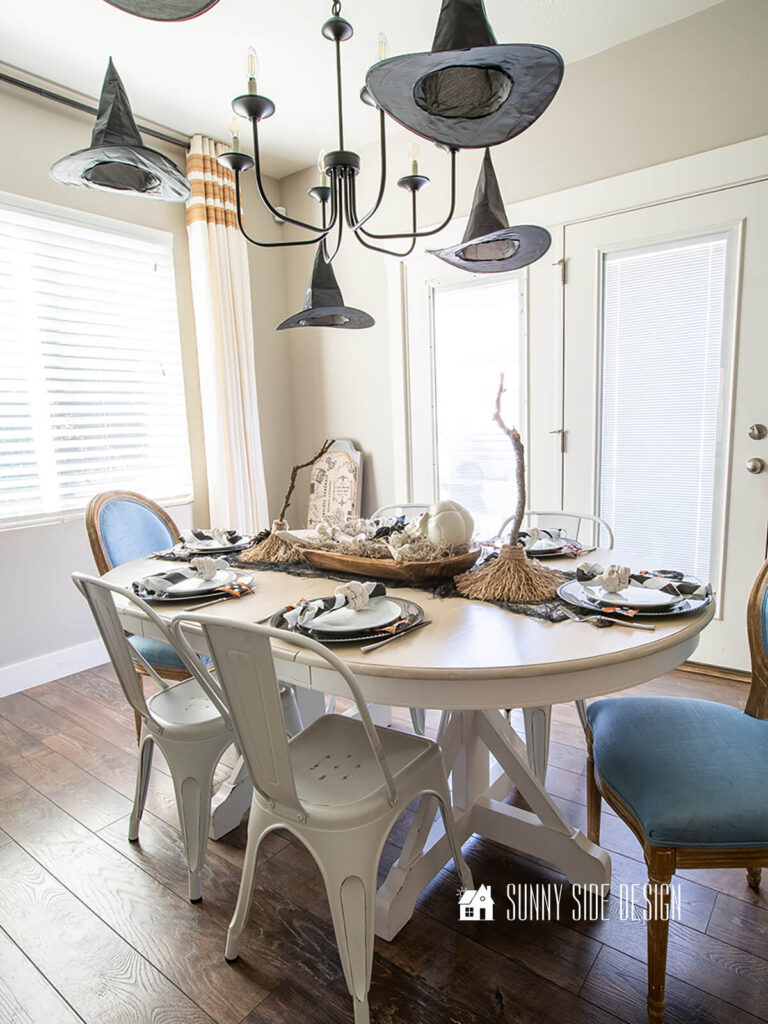Halloween decorating ideas: Floating witches hats are placed above the dining table. The dining table is decorated with a black creepy cloth table runner, dough bowl filled with spanish moss, skeleton bones and black birds. On each side of the dough bowl is a DIY witch broom.
