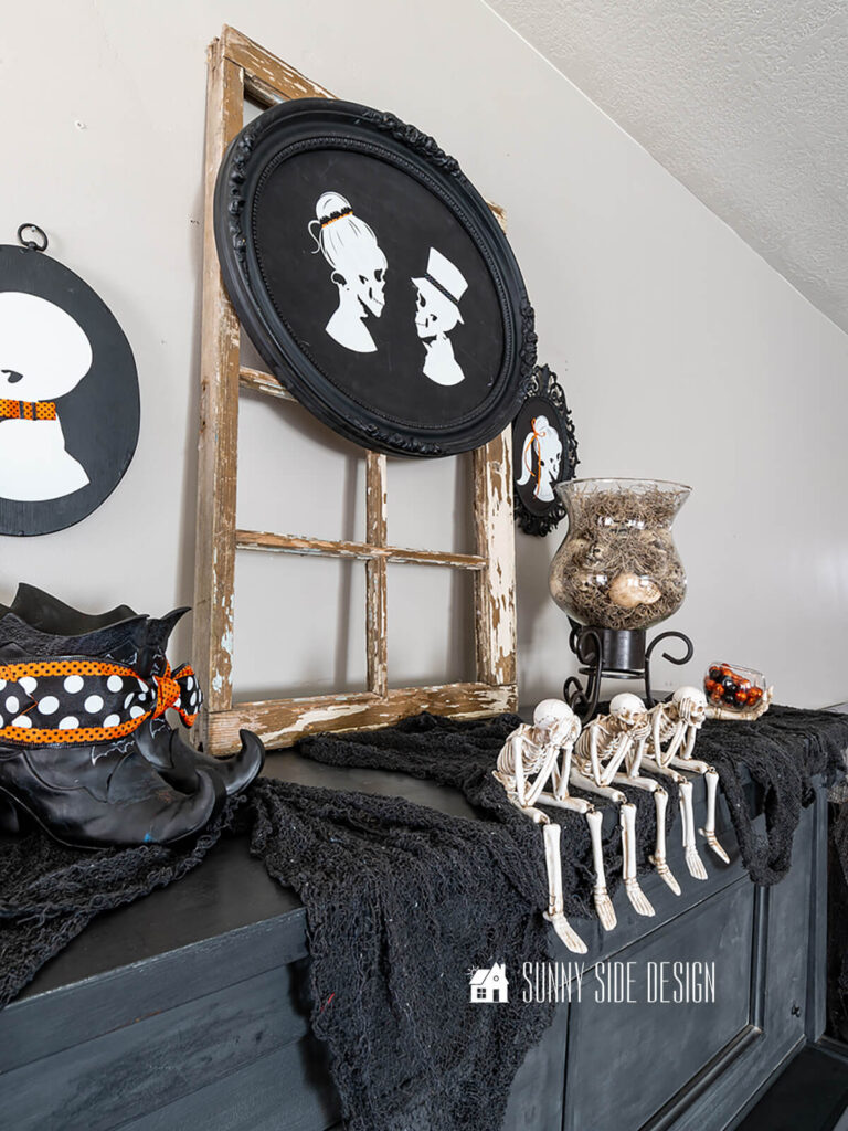 Antique heirloom piano is decorated for halloween with black creepy cloth, black vase, witches boots, glass hurricane filled with spanish moss and skulls, pumpkin and see no evil, hear no evil and speak no evil skeletons. Above the piano are DIY family skeleton portraits.