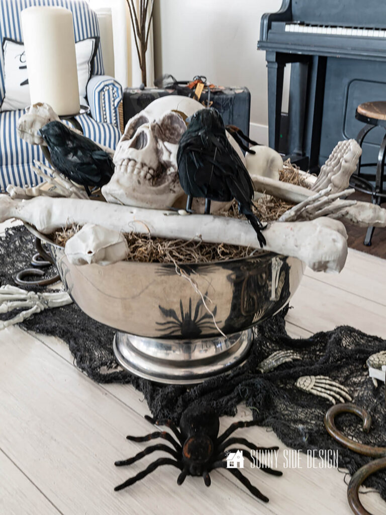 Coffee table decorated for Halloween with a black creepy cloth table runner, and a thrifted silver footed bowl filled with skeleton bones and black birds. Spiders are also placed on the coffee table.