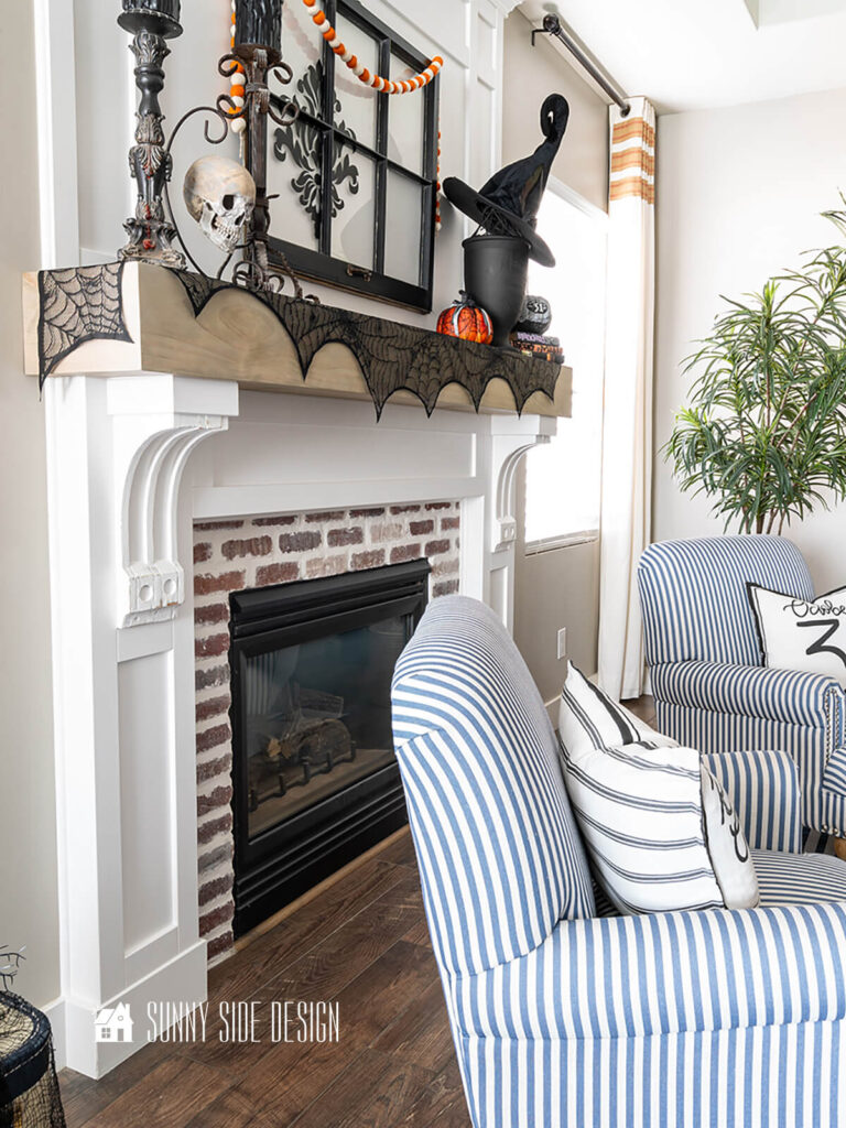 Fireplace mantle decorated for Halloween with a spider web mantle scarf, black vintage window embellished with a damask emblem, vintage candlesticks, witch hat and pumpkins.