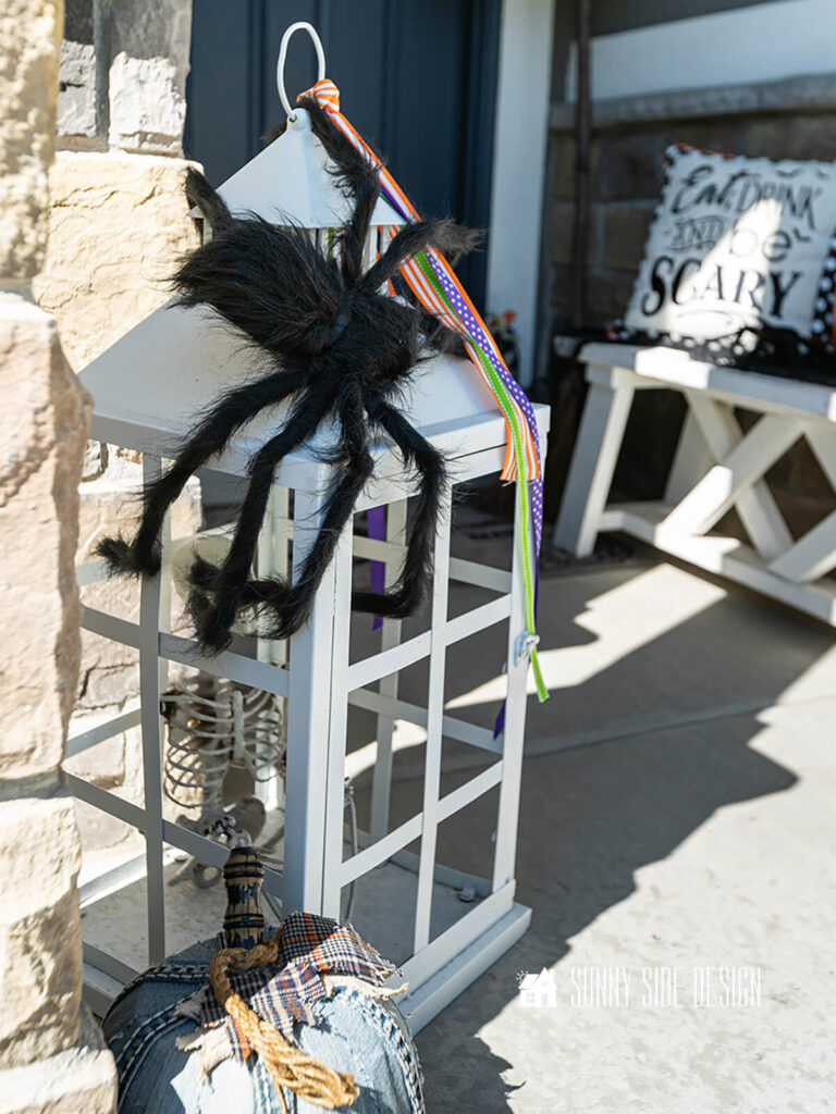White lantern embellished with halloween ribbons and a spooky hairy spider. You'll also find a skeleton caged in the lantern.