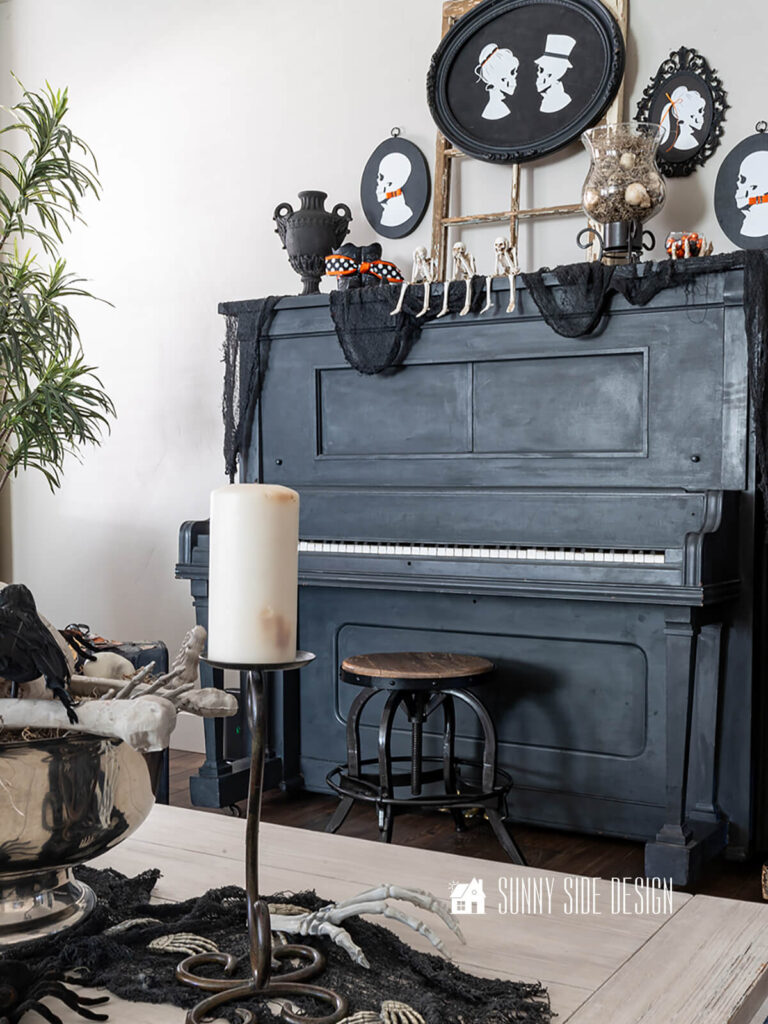 Antique heirloom piano is decorated for halloween with black creepy cloth, black vase, witches boots, glass hurricane filled with spanish moss and skulls, pumpkin and see no evil, hear no evil and speak no evil skeletons. Above the piano are DIY family skeleton portraits.