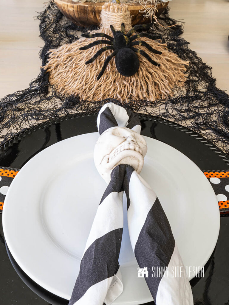 Halloween place setting with a black charger, a strip of halloween ribbon, white plate and DIY skull napkin rings with a black and white napkin.