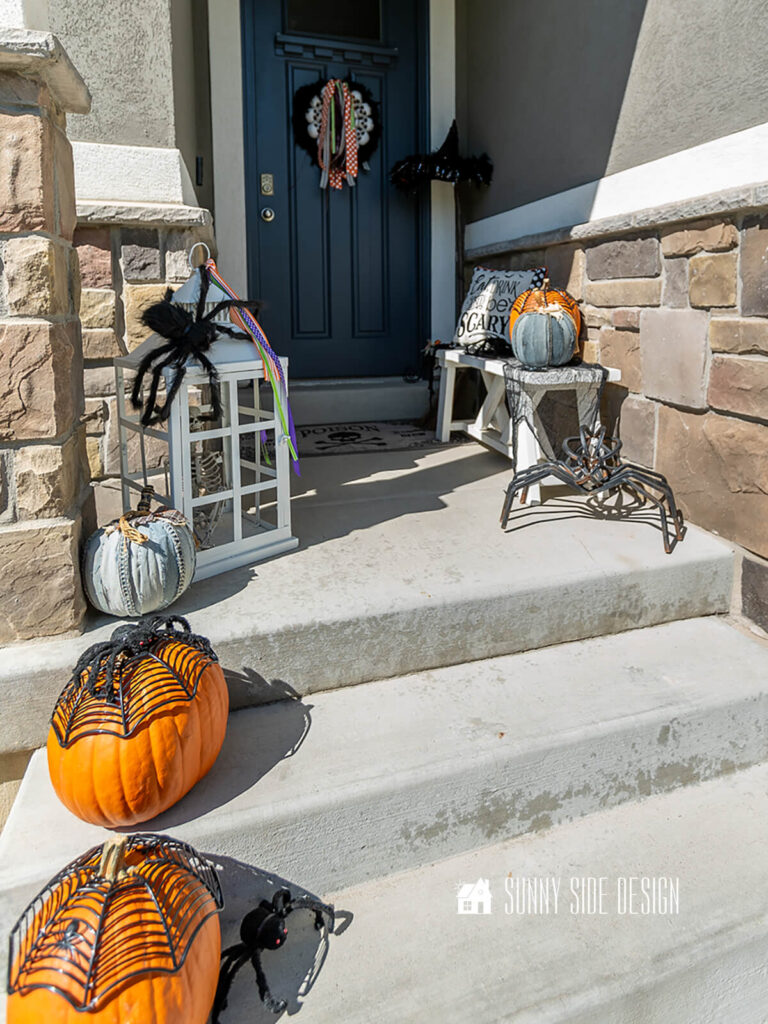 Front porch decorated for Halloween with spider web topped pumpkins, spooky lantern, bench with halloween pillow and pumpkins, witches broom and hat and a spooky black boa wreath embellished with skulls and ribbon.