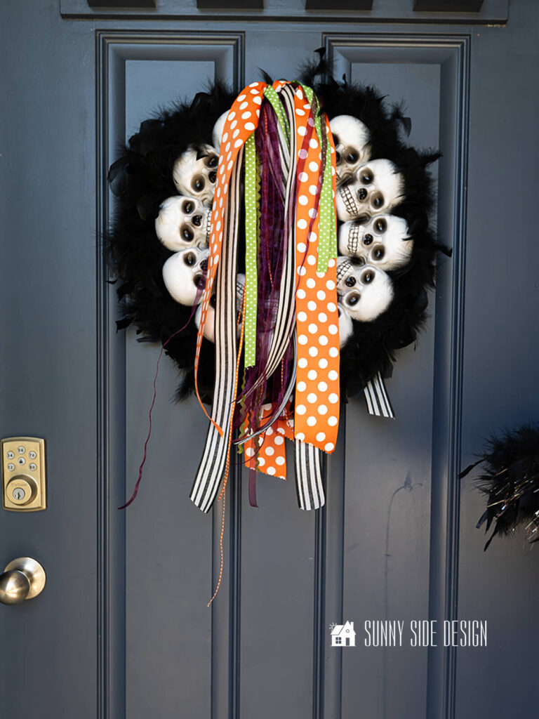 Black feather boa wreath embellished with skulls and halloween ribbon, hung on a navy blue front door.