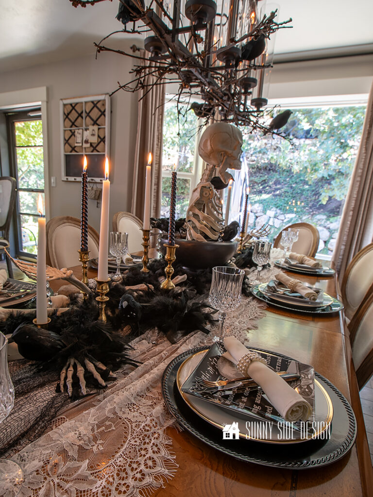Elegant Spooky Halloween Table Decor Ideas: Skeleton centerpiece in a black pedestal bowl, black and white candles in vintage candlesticks, lace and creepy cloth table runner, Black chargers with a gold rimmed plate, topped with a skeleton salad plate and crystal goblets.