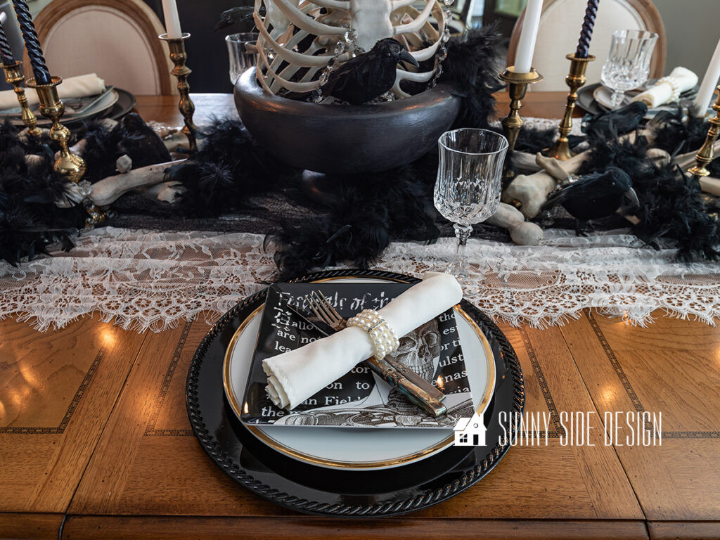Halloween table setting: Black charger topped with a thirfted gold rimmed white plate, and a black and white skeleton salad plate. Vintage silverware is set at each placesetting along with a white napkin and a rhinestone and pearl napkin ring.