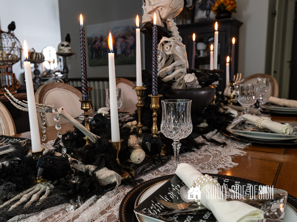 Elegant Spooky Halloween Table Decor Ideas: Skeleton centerpiece in a black pedestal bowl, black and white candles in vintage candlesticks, lace and creepy cloth table runner, Black chargers with a gold rimmed plate, topped with a skeleton salad plate and crystal goblets.
