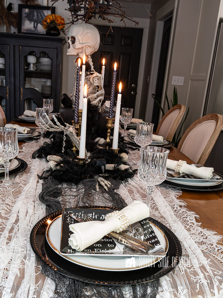 Elegant Spooky Halloween Table Decor Ideas: Skeleton centerpiece in a black pedestal bowl, black and white candles in vintage candlesticks, lace and creepy cloth table runner, Black chargers with a gold rimmed plate, topped with a skeleton salad plate and crystal goblets.