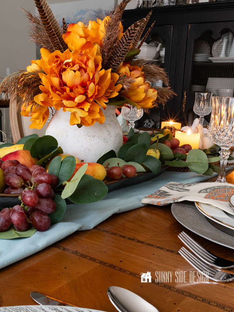 Simple Thanksgiving table decorations: light blue table runner with centerpiece of faux orange and pheasant flowers in a white rustic vase placed in a metal tray filled with assorted fresh fruit.