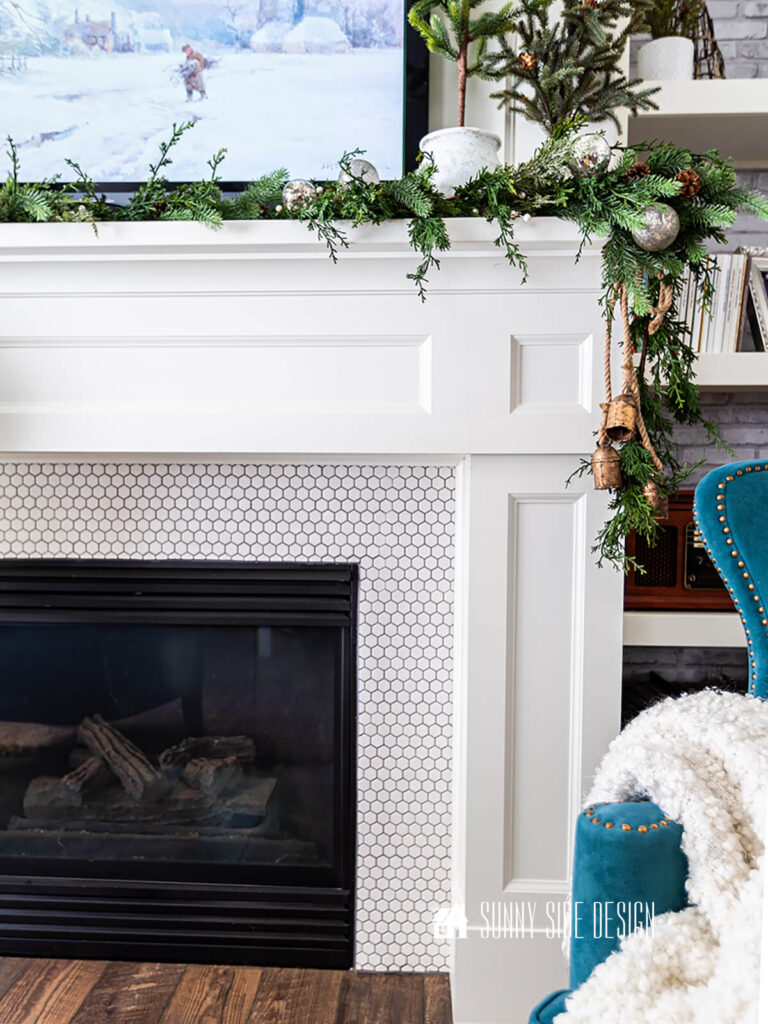 DIY brass Christmas bells are hung on the side of the mantle with greenery with a TV hung over the mantle with Winter art.
