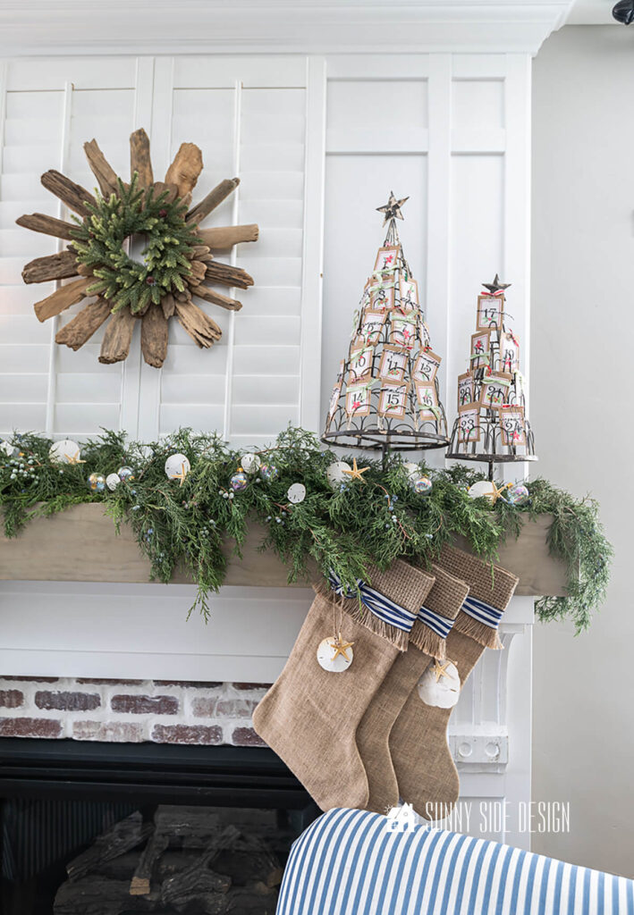 Fresh green juniper stems are placed on the mantle along with white and clear small glass ornaments and to complete the coastal Christmas mantle tiny starfish are added to the fresh greens. A tree advent calendar is placed on the right side and a drift wood wreath is hung on shutters over the mantle.