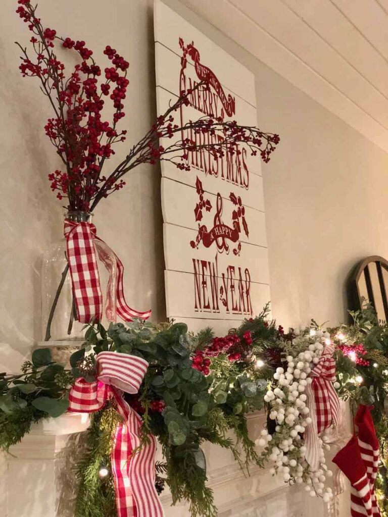 Traditional red, white and green Christmas decor, a variety of greens are layered on the mantle with twinkle lights and red and white berries. A vase is also filled with red berry stems tied with red and white ginham ribbon. Red and white Christmas plaque is placed above the mantle.