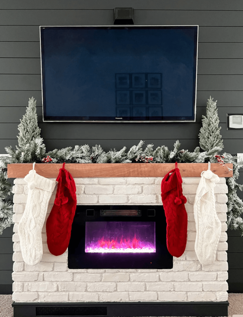 Christmas Decorating Ideas for Mantle with a TV. Simple flocked garland is placed on the mantle along with two flocked mini Christmas trees and 4 red and white stockings are hung from the mantle.