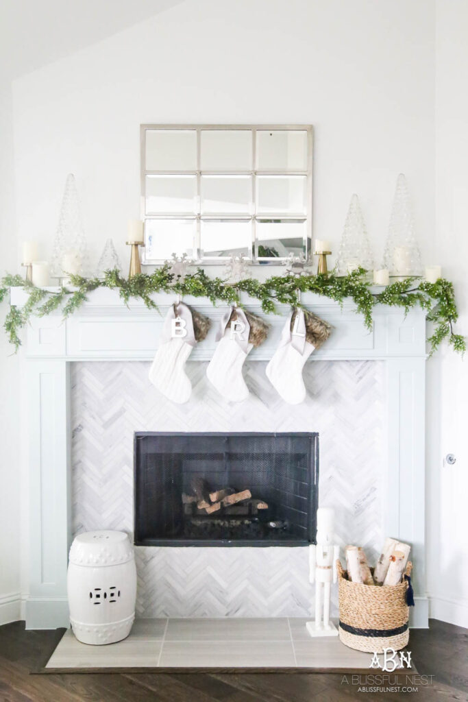 Winter white Christmas decorating ideas for a mantle with a simple green garland, white Christmas table top trees, white candles and white stockings hung from the mantle.