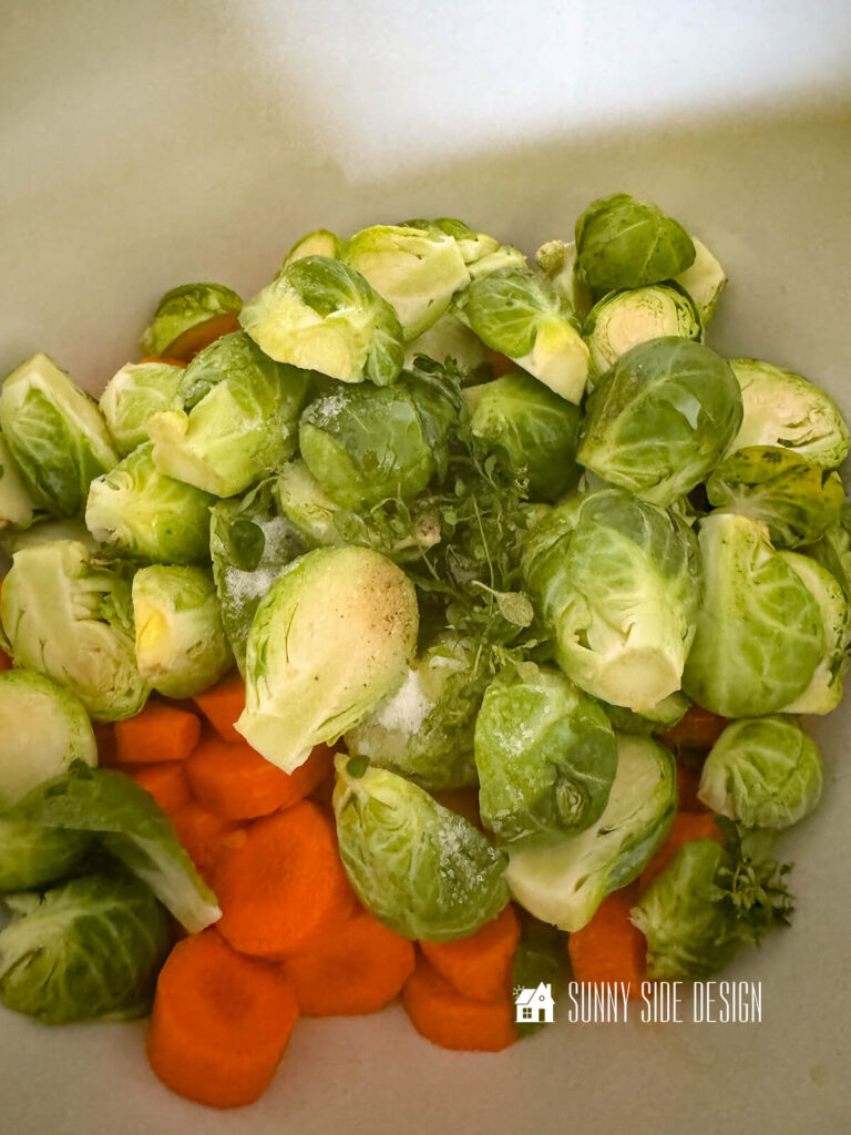 Halved brussel sprouts, sliced carrots, seasonings and olive oil in a white bowl.