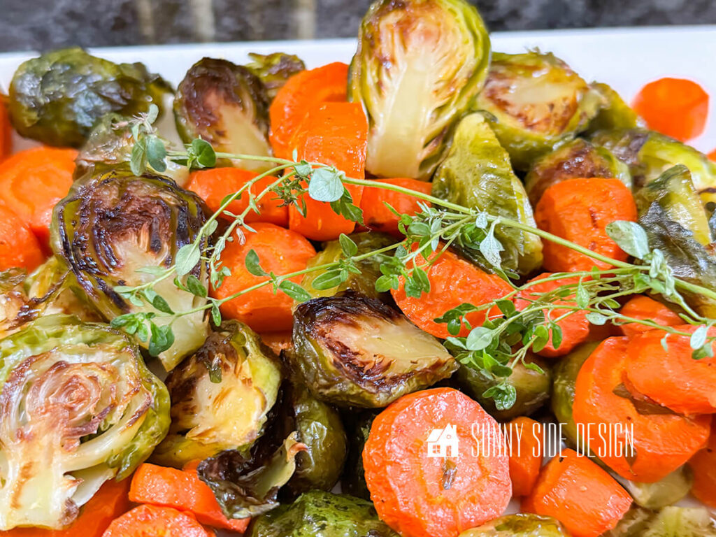 Crispy, browned roasted carrots and brussel sprouts, garnished with fresh thyme on a platter.
