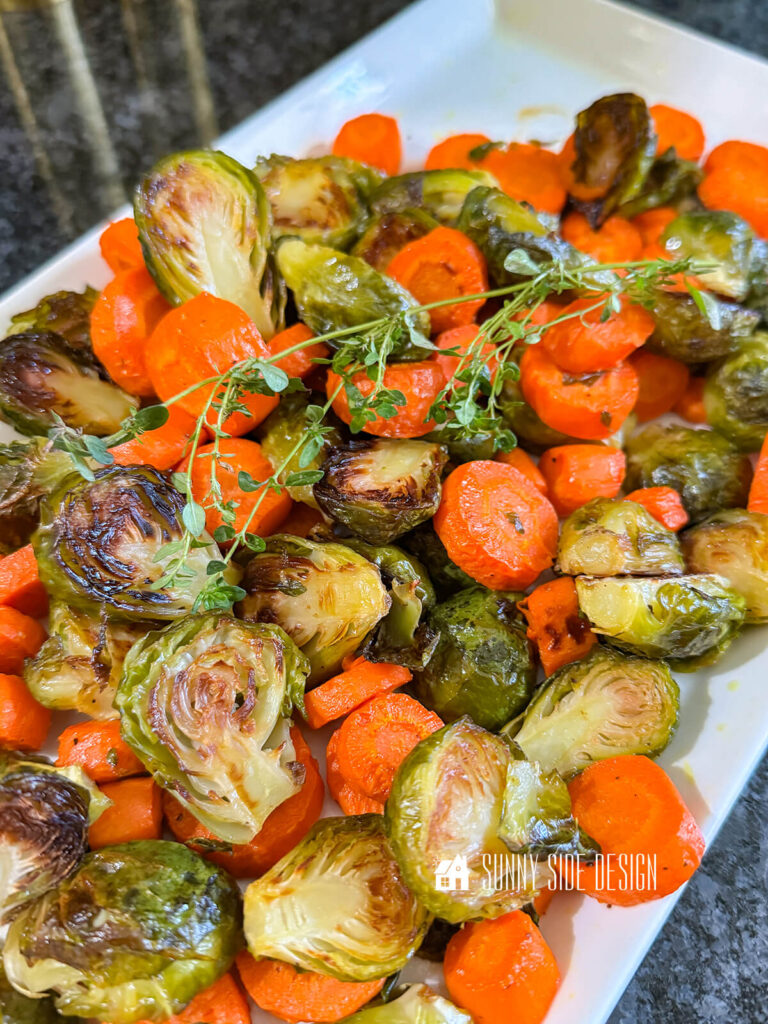 Crispy, browned roasted carrots and brussel sprouts, garnished with fresh thyme on a platter.