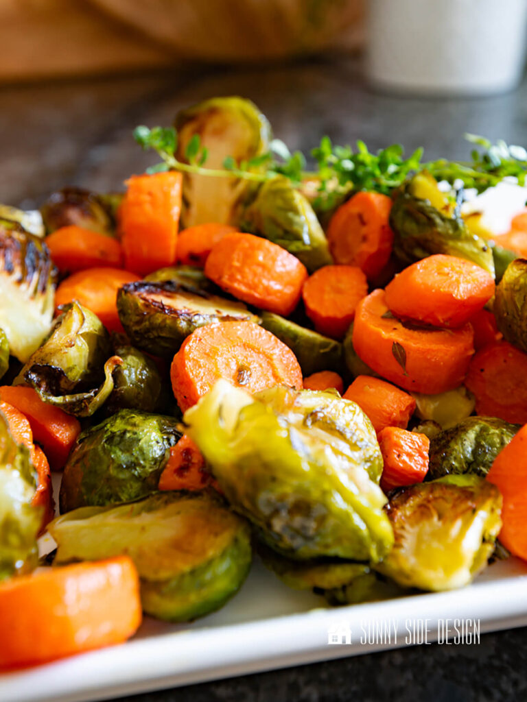 Crispy, browned roasted carrots and brussel sprouts, garnished with fresh thyme on a platter.