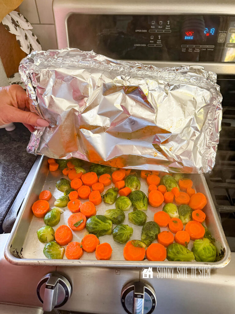 Woman pulls back the foil on a sheet pan with sliced carrots and halved brussel sprouts.