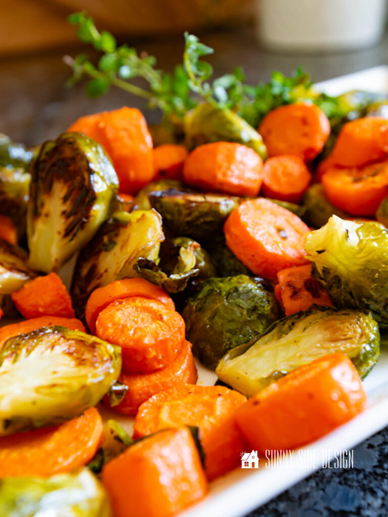 Crispy, browned roasted carrots and brussel sprouts, garnished with fresh thyme on a platter.