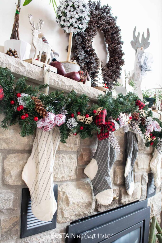 Woodsy and rustic Christmas mantle decorated with green garland, red berries, pinecones and stockings. A pinecone wreath is placed above the fireplace along with assorted deer.
