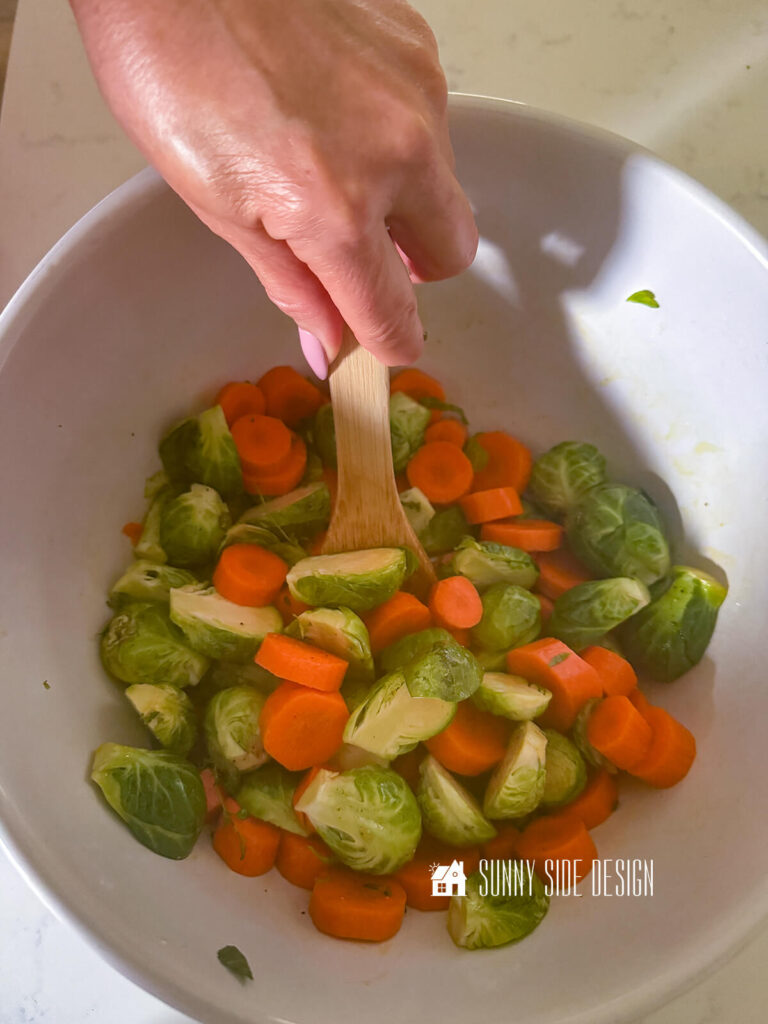 Woma, tosses brussel sprouts, carrots, olive oil and seasonings in a bowl with a wooden spoon.