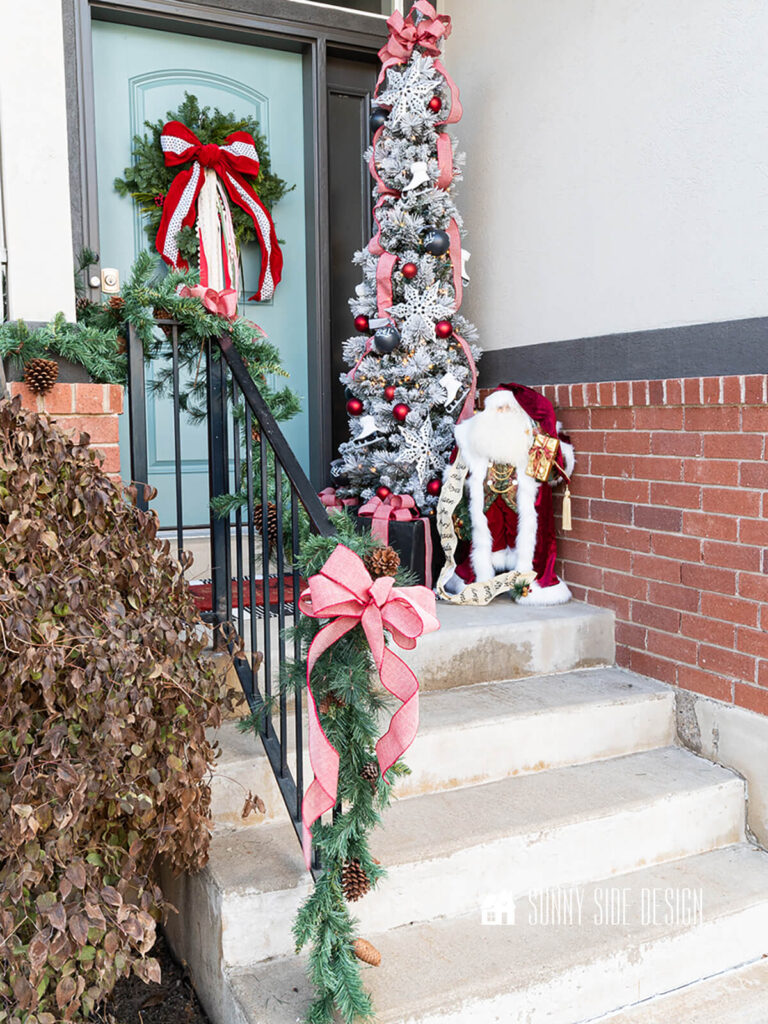 Small Christmas porch styled with a fresh wreath and festive ribbons, slim flocked tree with red ball ornaments, ice skate ornaments, black noel ornaments and red bow topper, garland and red bows on the railing and festive Santa.