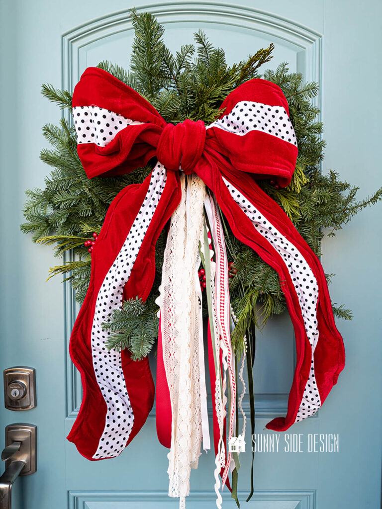 Fresh wreath on a blue front door embellished with red berries and a large red velvet bow and assorted red, green and white ribbons.