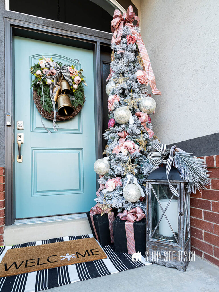 Small Christmas porch with a grapevine wreath embellished with faux greens, blush peonies, black and white ribbon and large faux brass bells. Slim pencil flocked tree with white and blush ornaments and blush floral bouquets. Faux presents and a large lantern embellished with greenery and black and white ribbon.