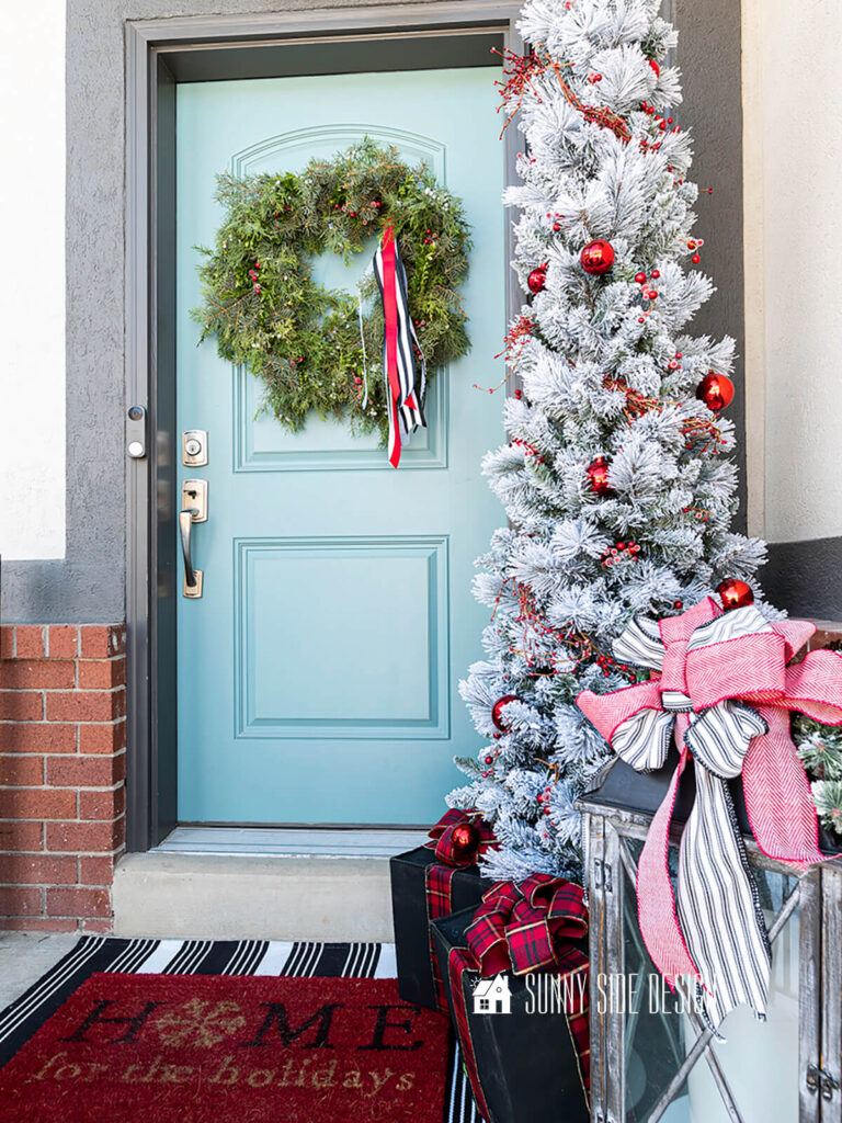 Small Christmas porch styled with a fresh wreath accented with berries and red, white and black ribbon. Flocked pencil tree decorated with red berries and red ball ornaments, faux gift boxes at the base and a lantern embellished with red, black and white ribbon and greenery.