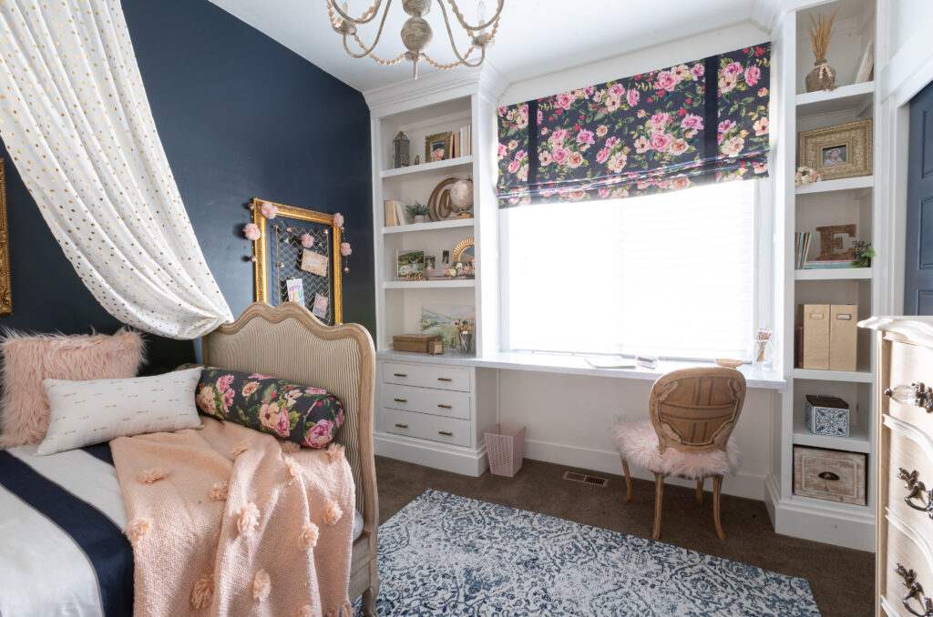 Navy blue accents wall, built-in bookshelves with desk area around the window with navy floral roman shades, french inspired daybed with a bed canopy, boho chandelier and a blue and white patterned rug.