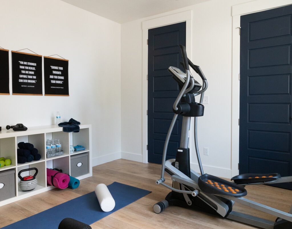Stephanie's Home gym with white walls, navy blue doors, and french oak laminate floors. Inspirational framed quotes on the wall above an Ikea cabinet for gym supplies, elliptical, yoga mat and foam roller.