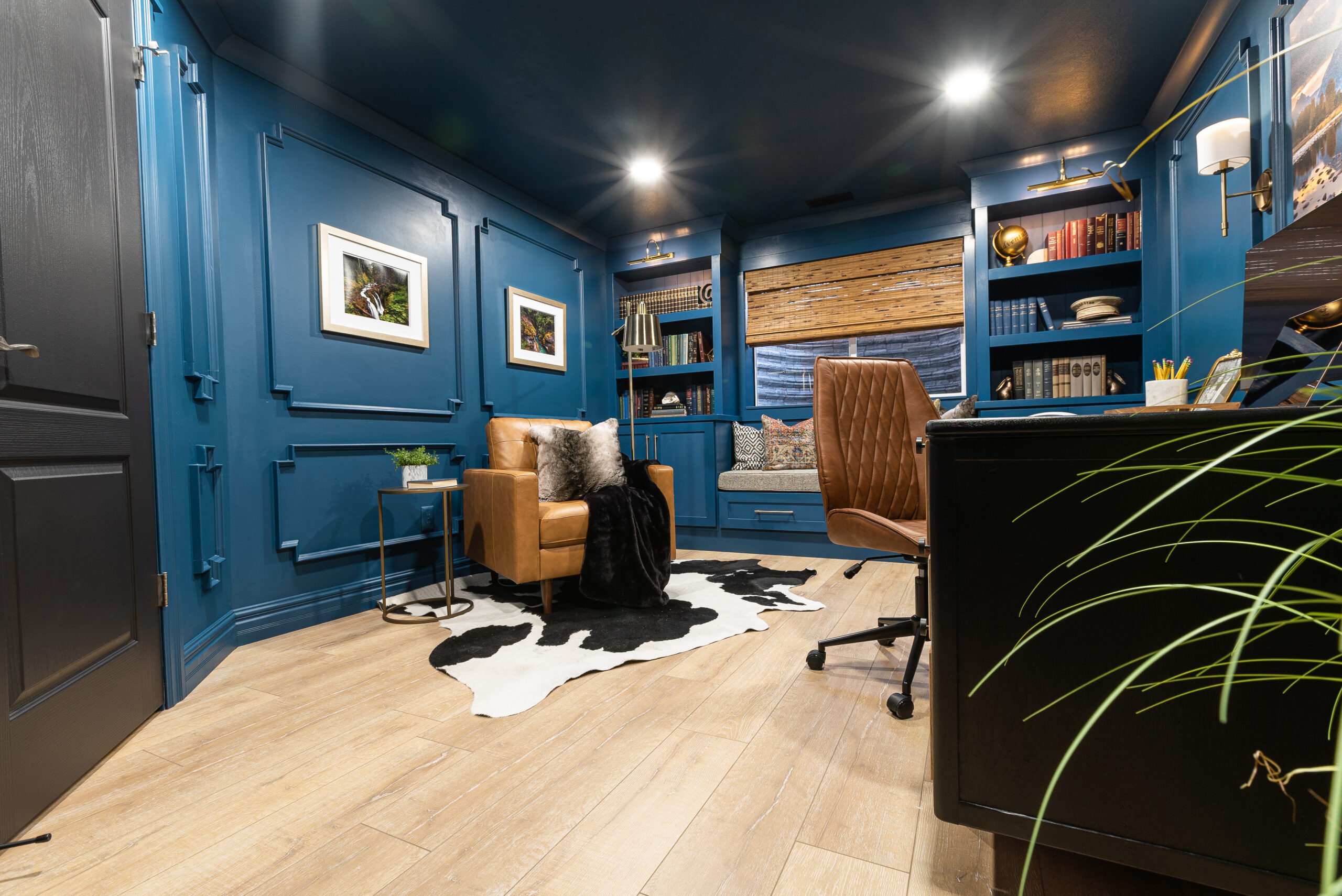 Moody home office with library in a once unfinished basement bedroom space. Now with dark blue box moulding walls, french oak laminate floor, modern desk with leather chair, leather reading chair and built-in bookshelves with a window seat.
