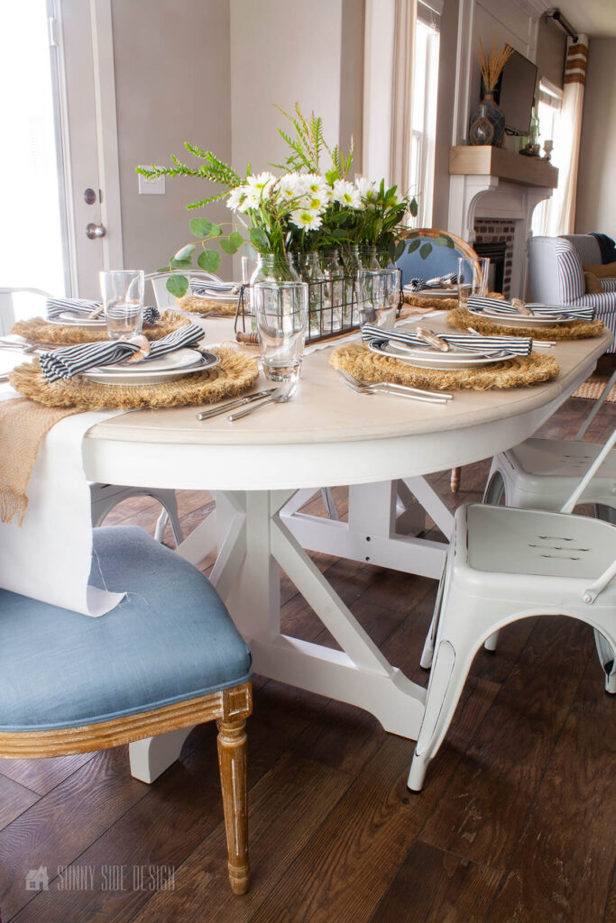 Just off the living room is the dining area with a refinished oak table with a coastal look. There are 2 upholstered blue chairs and 4 white farmhouse metal chairs. Table decorated with fresh flower and table settings.
