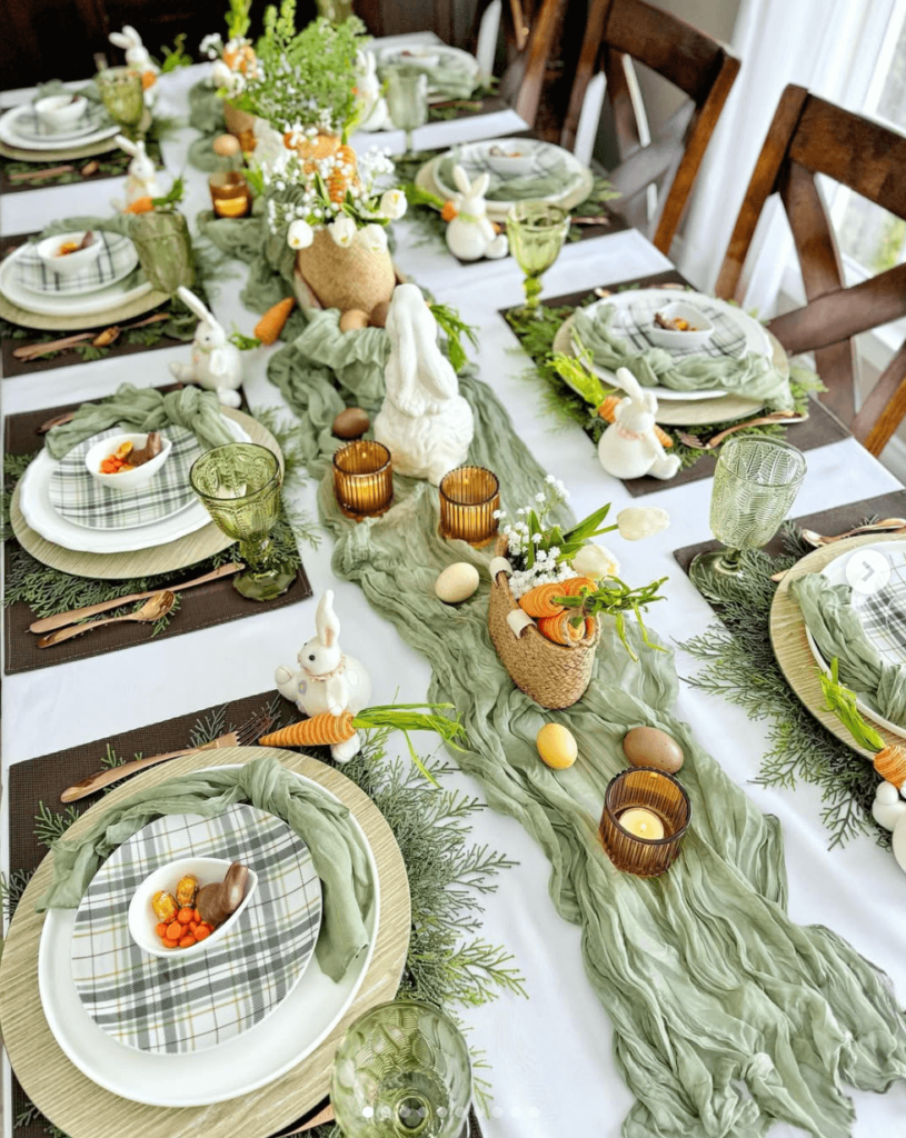 Easter tablescape with green gauze table runner and knotted napkins, plaid sage green plates on natural wood chargers, the centerpiece features ceramic bunnies, eggs, mini baskets filled with carrots and lily of the valley and amber votive candles.