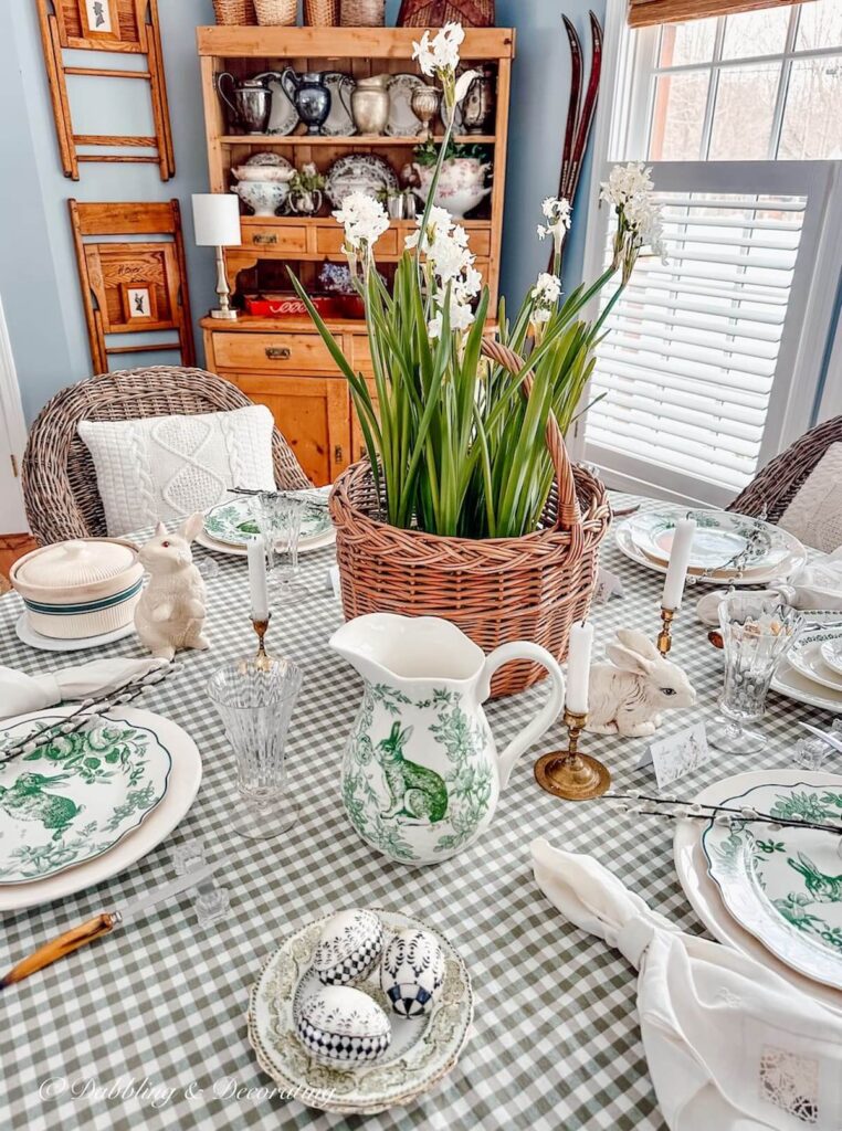 Spring table decor ideas with a green gingham table cloth, vintage linen napkins, green and white bunny plates, and paper whited in a basket as the centerpiece.