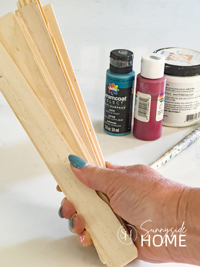 Woman holds a stack of wood paint stir sticks with red, white and blue paint in the background.