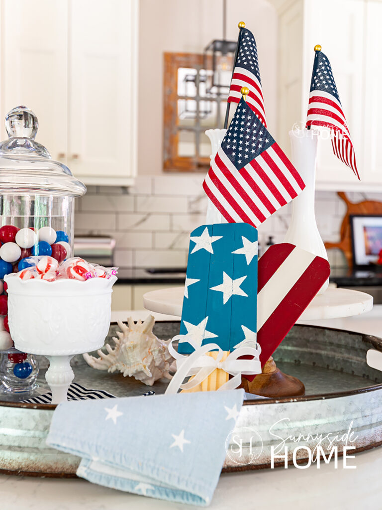 Blue and white star, red and white stripe painted patriotic decor using paint stir sticks. Styled with an apothecary jar filled with red white and blue gumballs, white footed dish with peppermints and mini flags in bud vases.