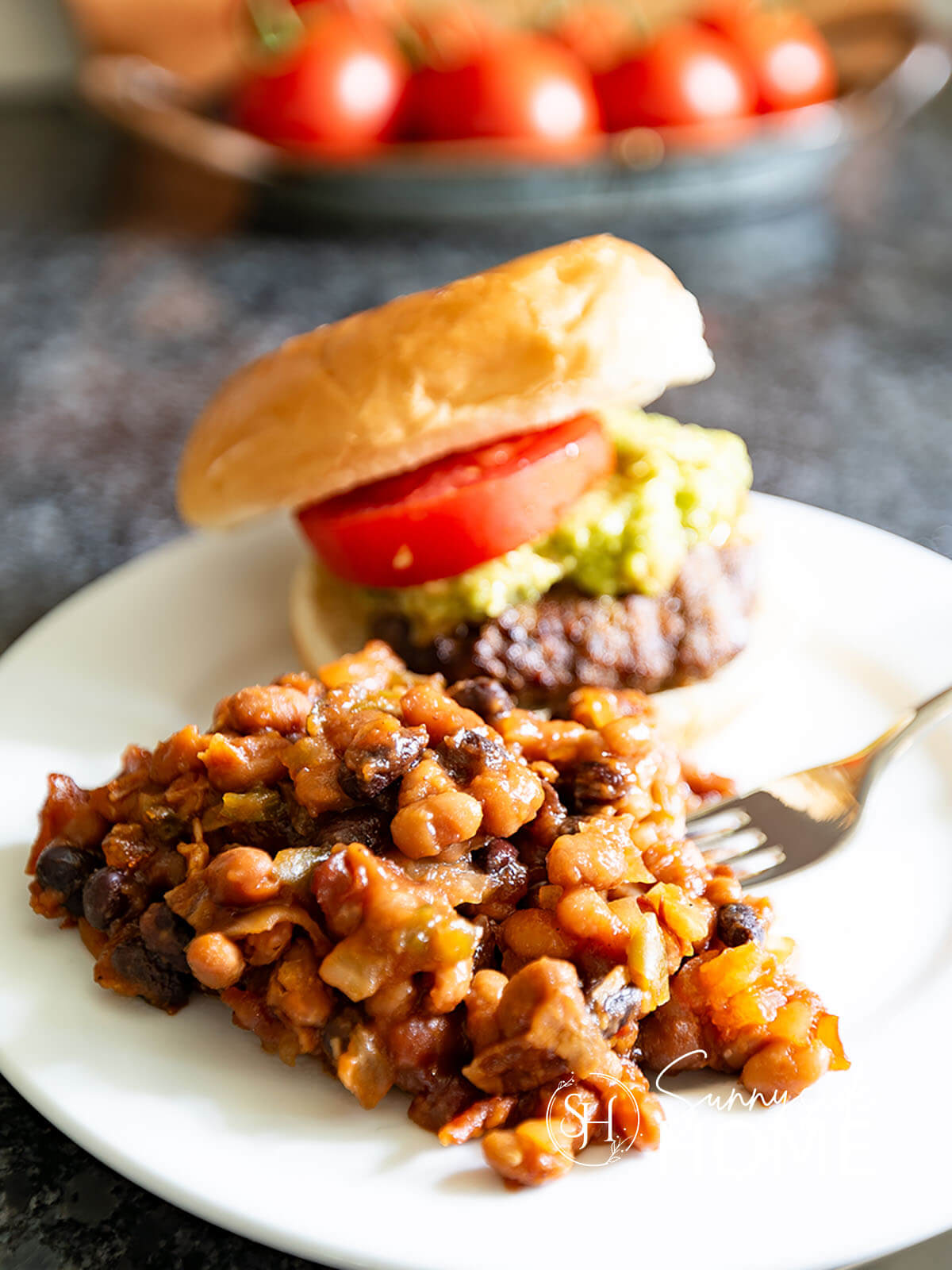 Homemade Maple Baked Beans served with a hamburger topped with guacamole and tomato.