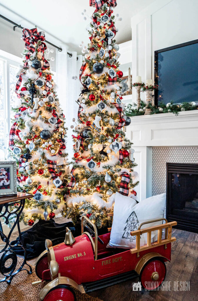 A pair of decorated Christmas trees by the fireplace with an old red pedal fire truck.