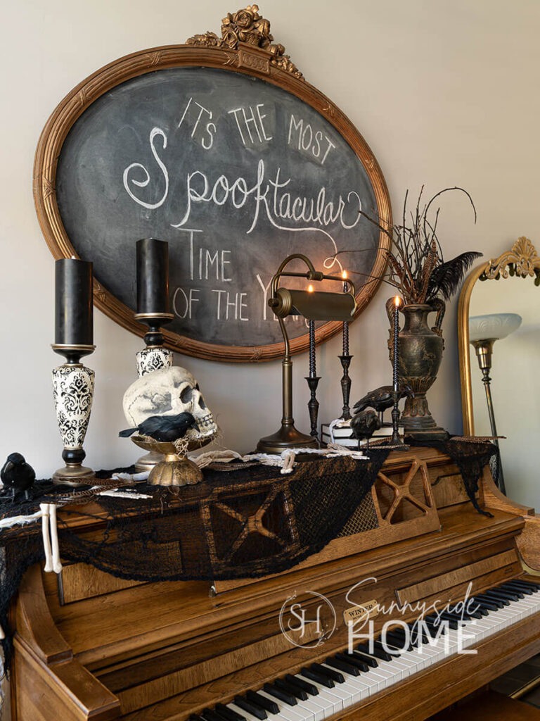 Piano is decorated for halloween with black candles, creepy cloth, skull with a black bird, more birds on book stacks and a black vase filled with twigs, feathers etc, A chalkboard sign is hung over the piano witha spooktacular saying.