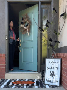Woman at door with Halloween wreath and a tree branch decorating for Halloween with moss and black birds.
