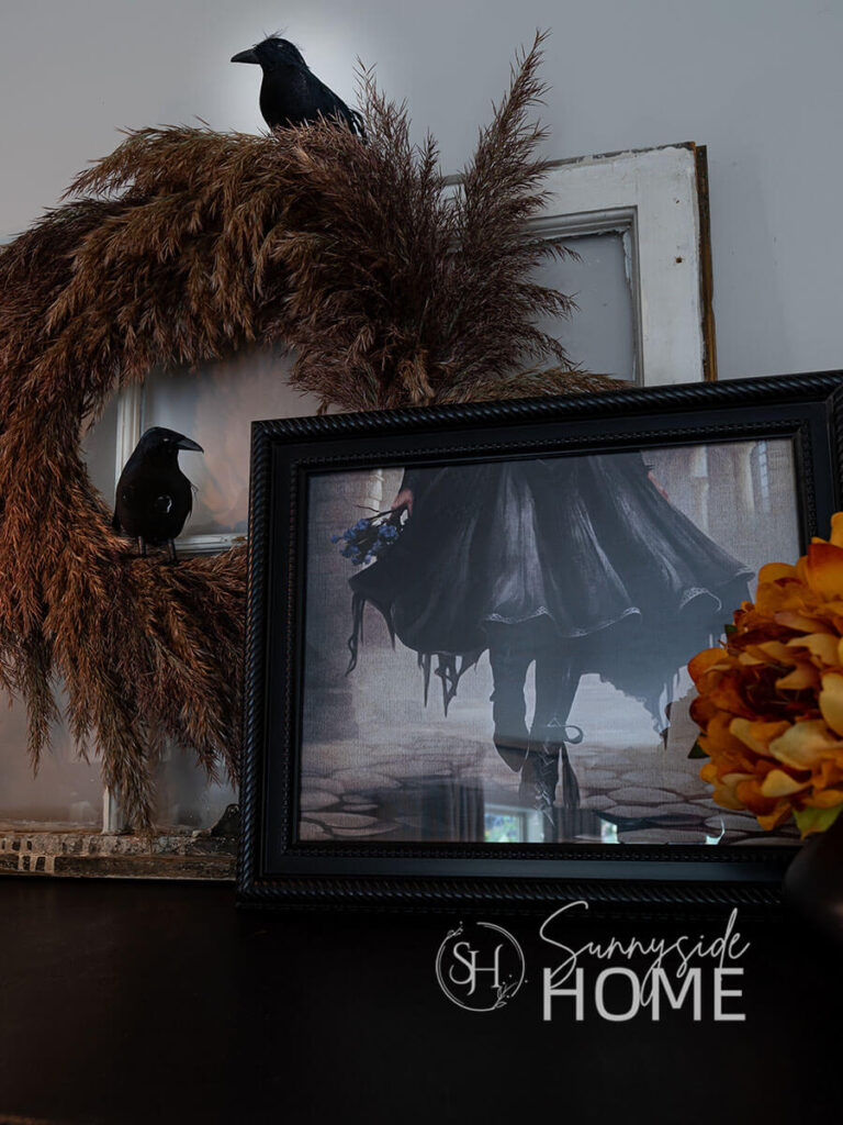 The hutch is decorated for halloween with framedHalloween print, pampas grass wreath adorned with black birds and orange mums.