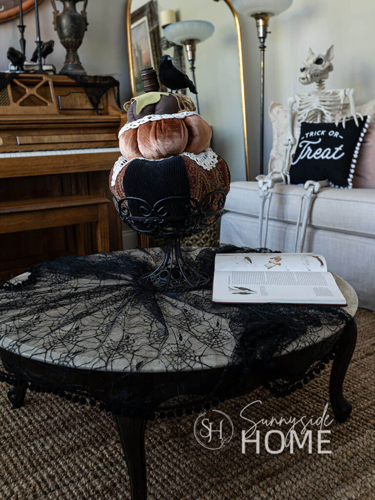 Decorating for Halloween, the coffee table is styled with black gothic lace, and a velvet pumpkin stack in a iron footed bowl, a book with images of birds and a skeleton sitting on the sofa with a trick or treat pillow.