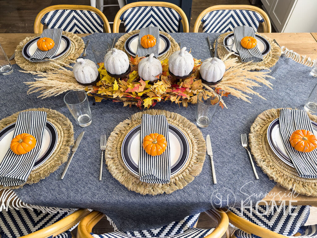 Simple and rustic fall table with a navy blue and white throw blanket as a tablecloth on a rustic wood table, placesettings feature rustic straw placemat, blue rimmed dinner plate topped with a blue and white napkin and an orange mini pumpkin. The centerpiece features a thrifted candle stand embellished with fall leaves and grasses and stone look mini pumpkins.