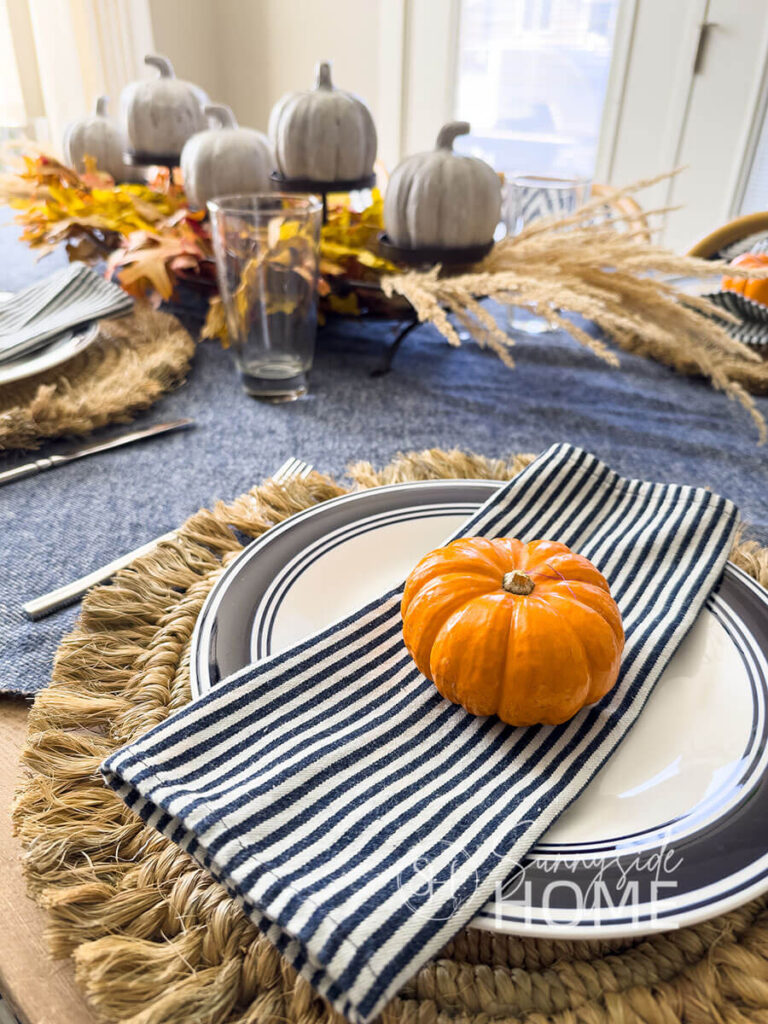 Simple and rustic fall table with a navy blue and white throw blanket as a tablecloth on a rustic wood table, placesettings feature rustic straw placemat, blue rimmed dinner plate topped with a blue and white napkin and an orange mini pumpkin. The centerpiece features a thrifted candle stand embellished with fall leaves and grasses and stone look mini pumpkins.